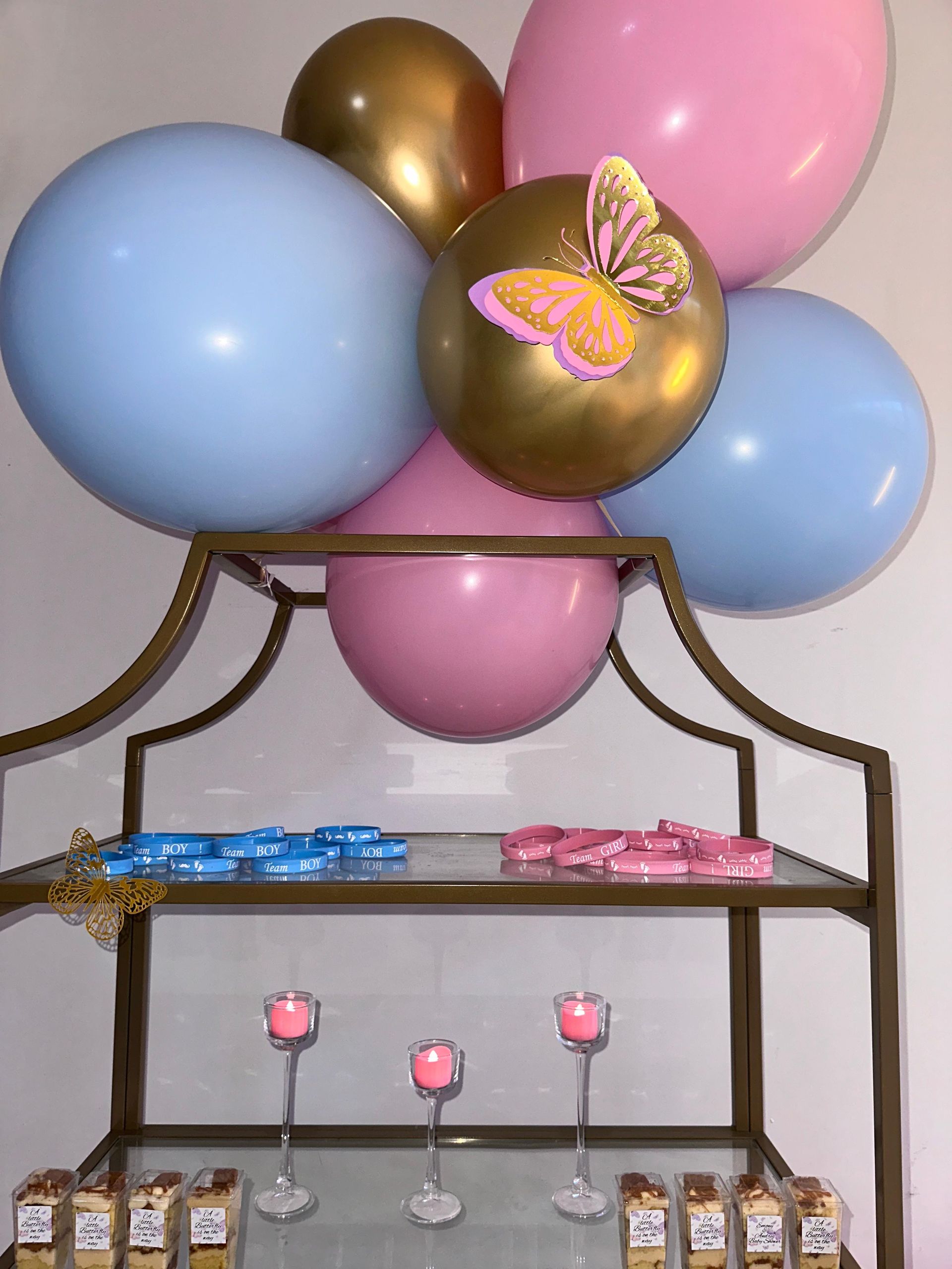 A party display features pink, blue, and gold balloons with a butterfly decal, tiered glass shelves, and candle holders.