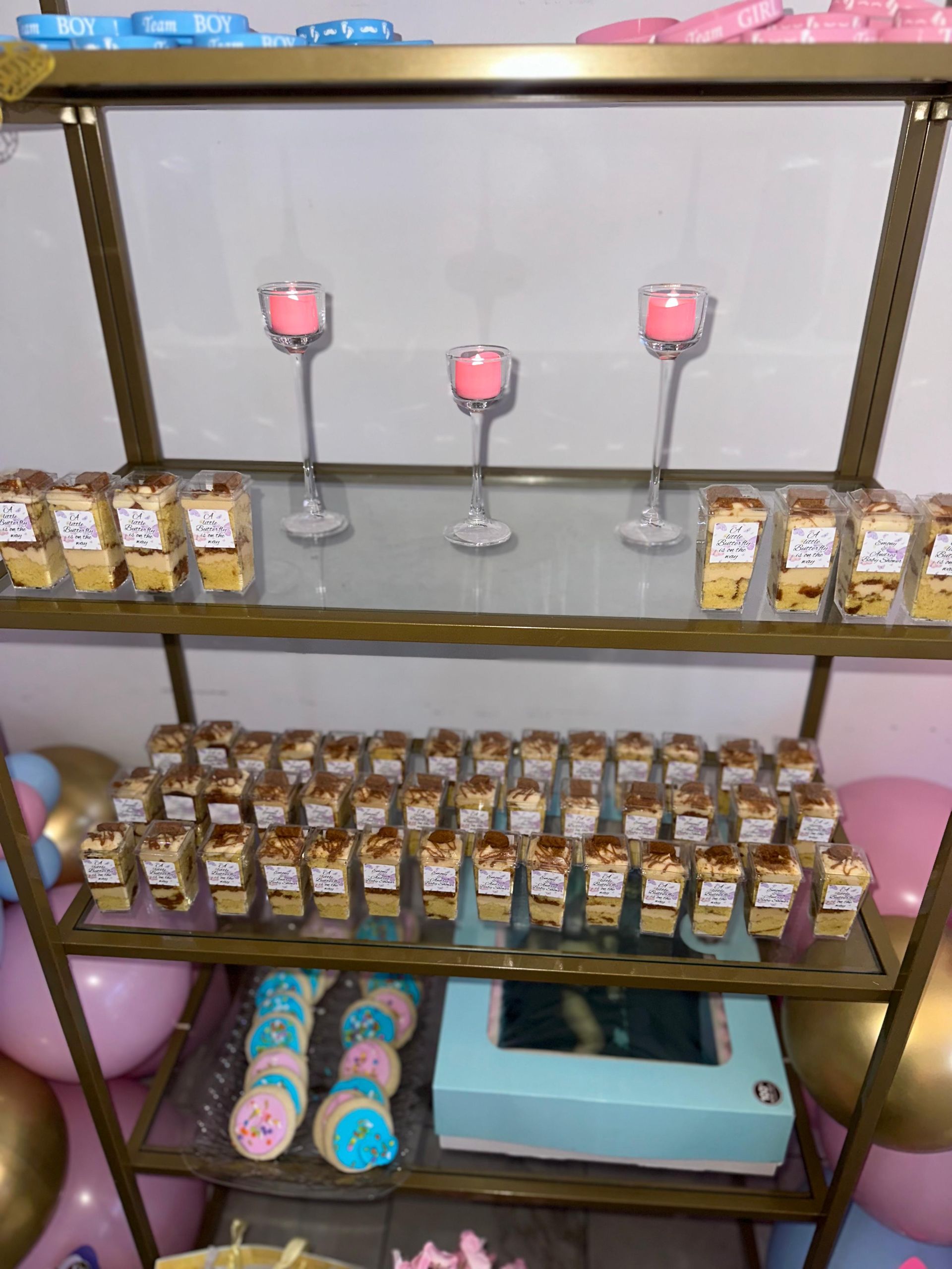 A gold display shelf holds small dessert cups, three pink candles, and blue-and-pink cake pops above balloons.