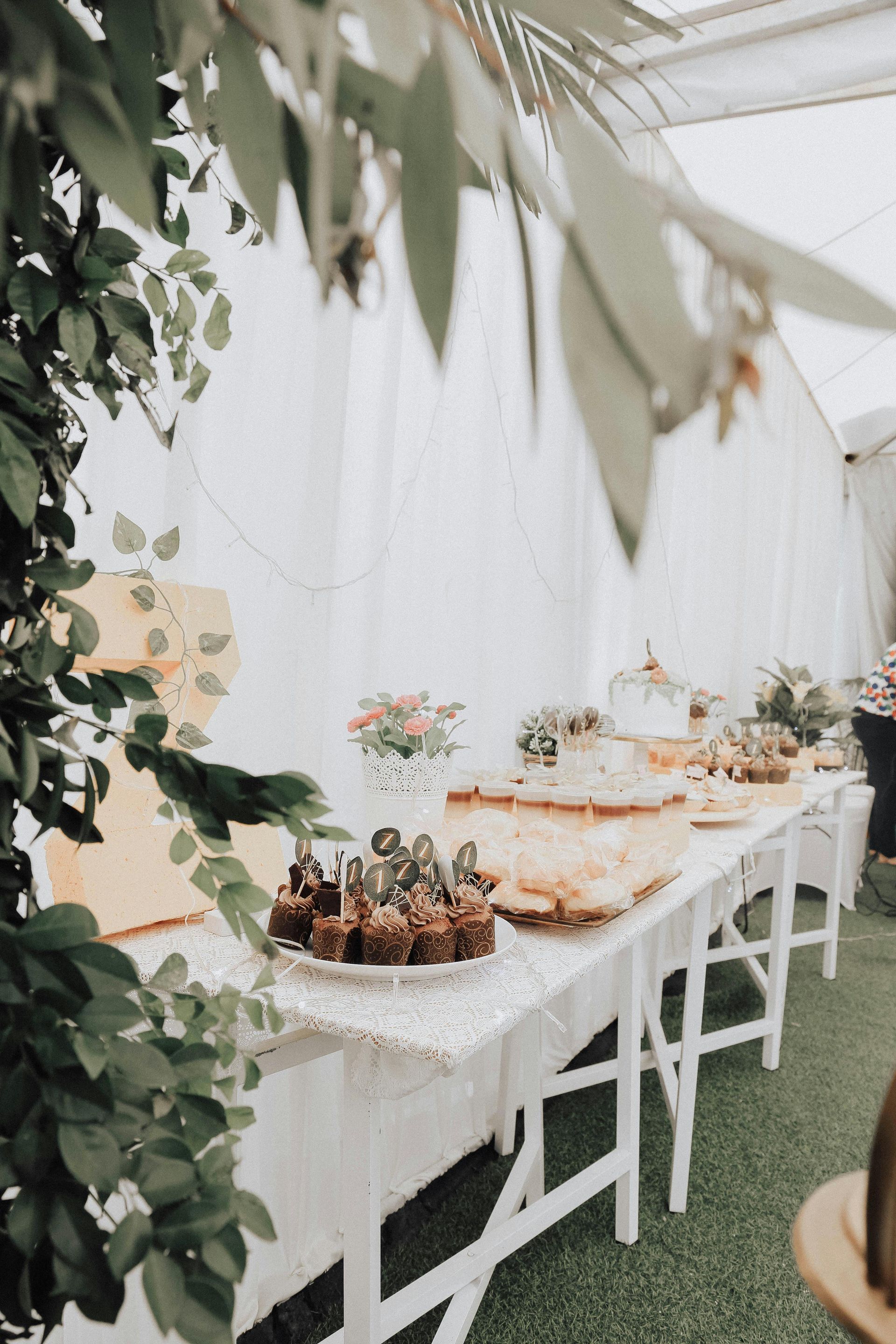 Long white table with desserts and flowers, set inside a marquee with greenery.