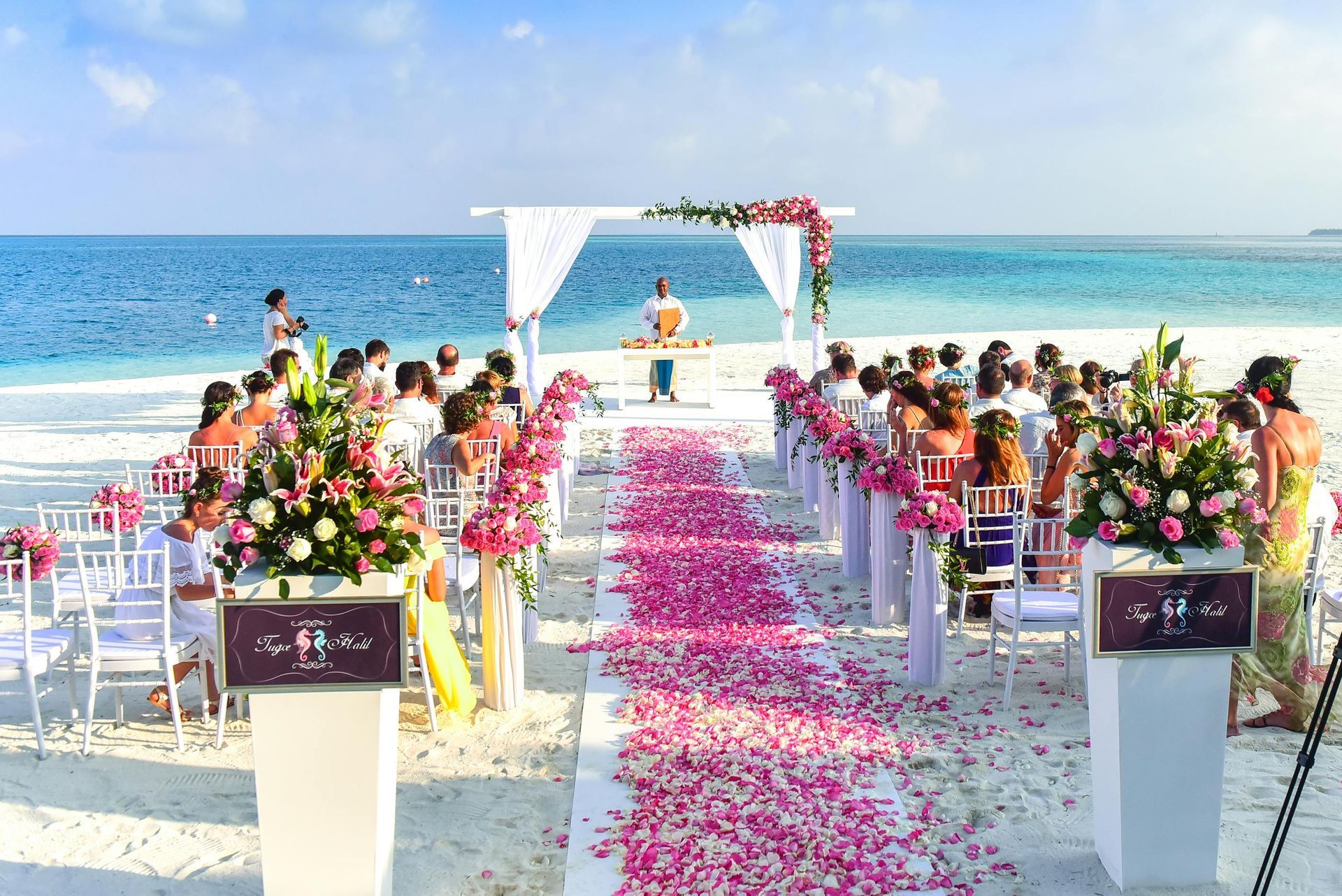 Beach wedding ceremony with pink flowers, white chairs, and ocean view.
