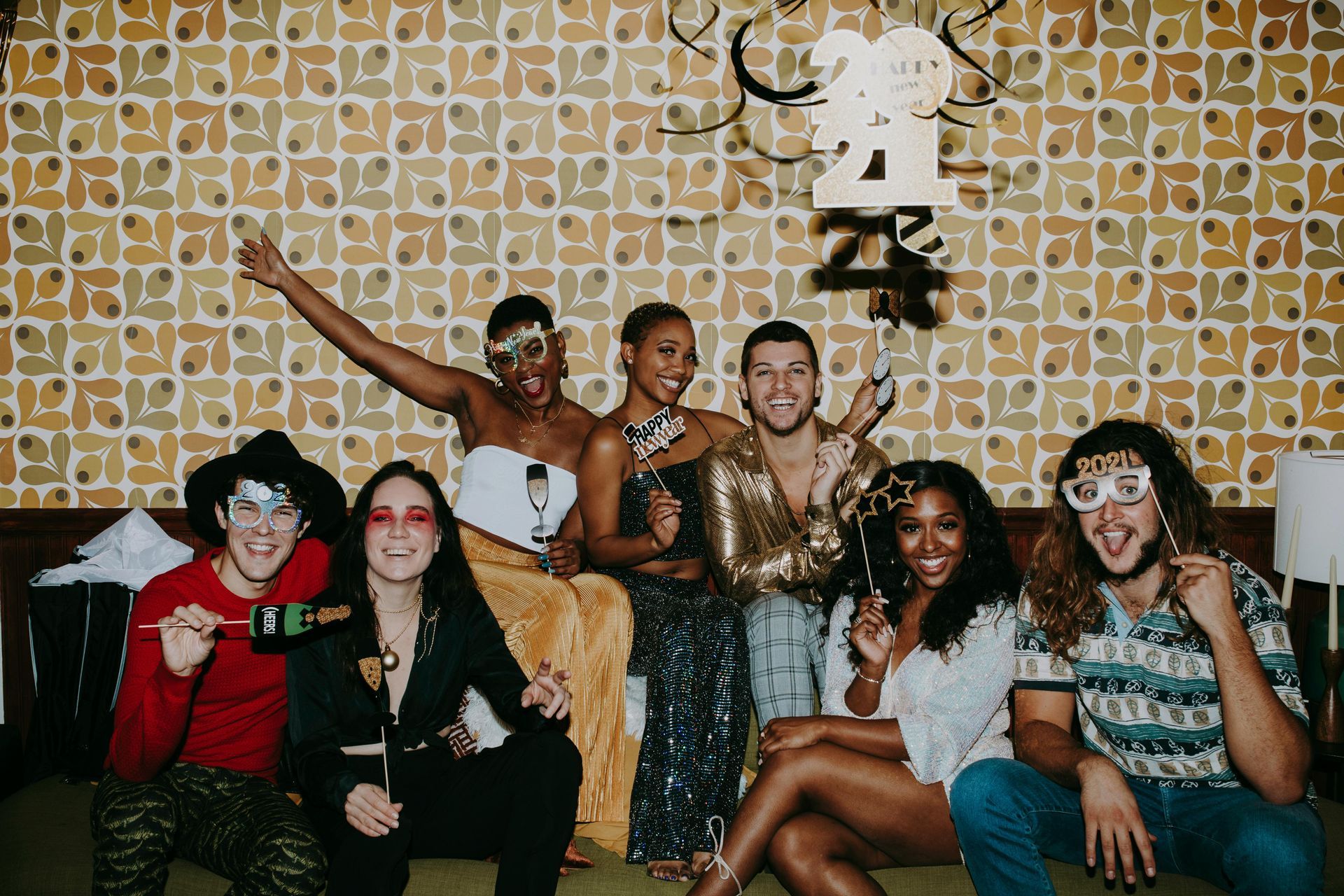 Group of friends celebrating, posing with drinks, fun outfits, in front of patterned wall, party vibe.