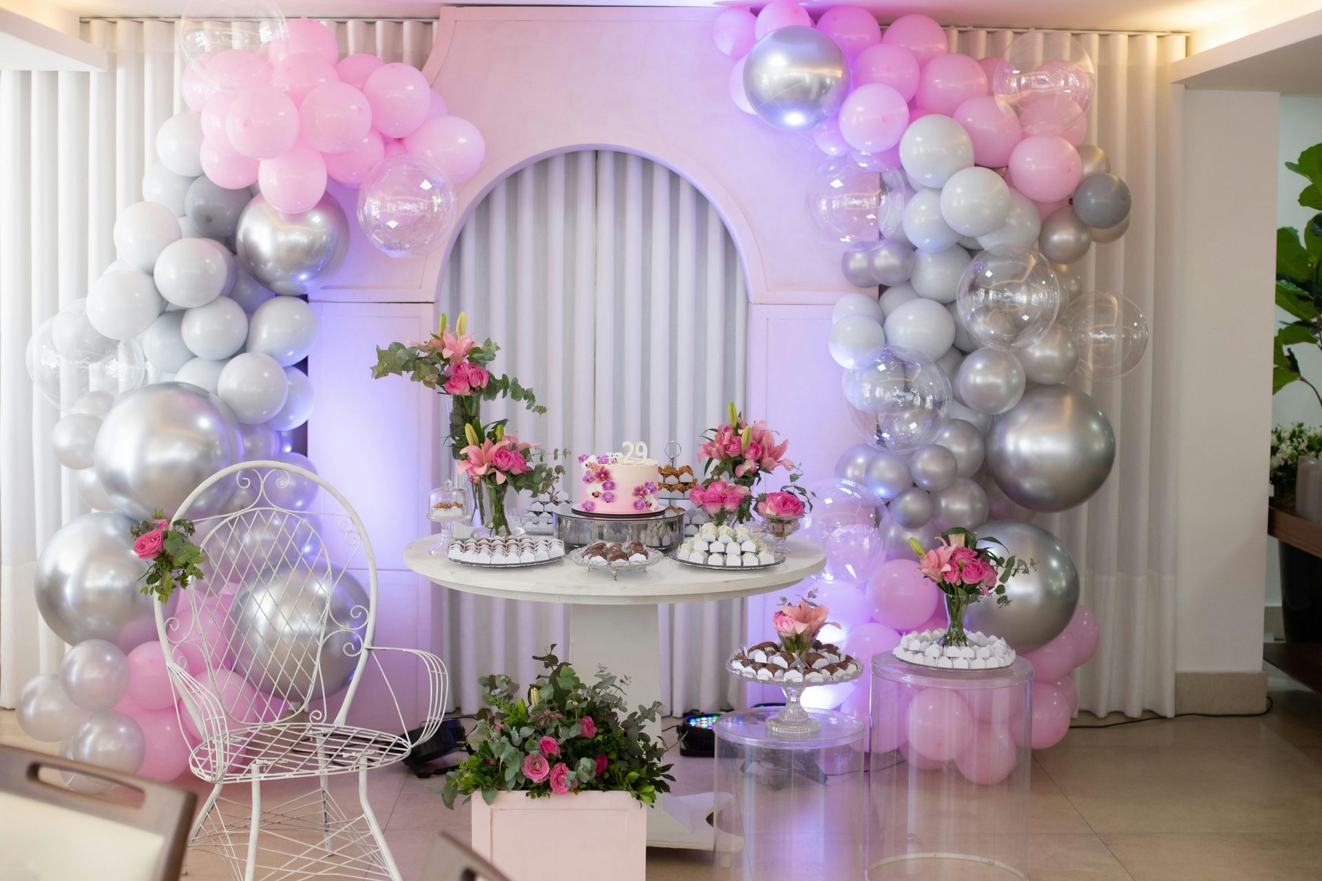 Birthday party setup with pink, silver, and white balloons around a table with cake and flowers.