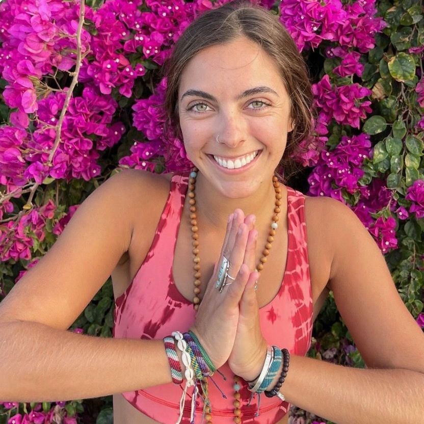 A woman is smiling in front of a bush of purple flowers.