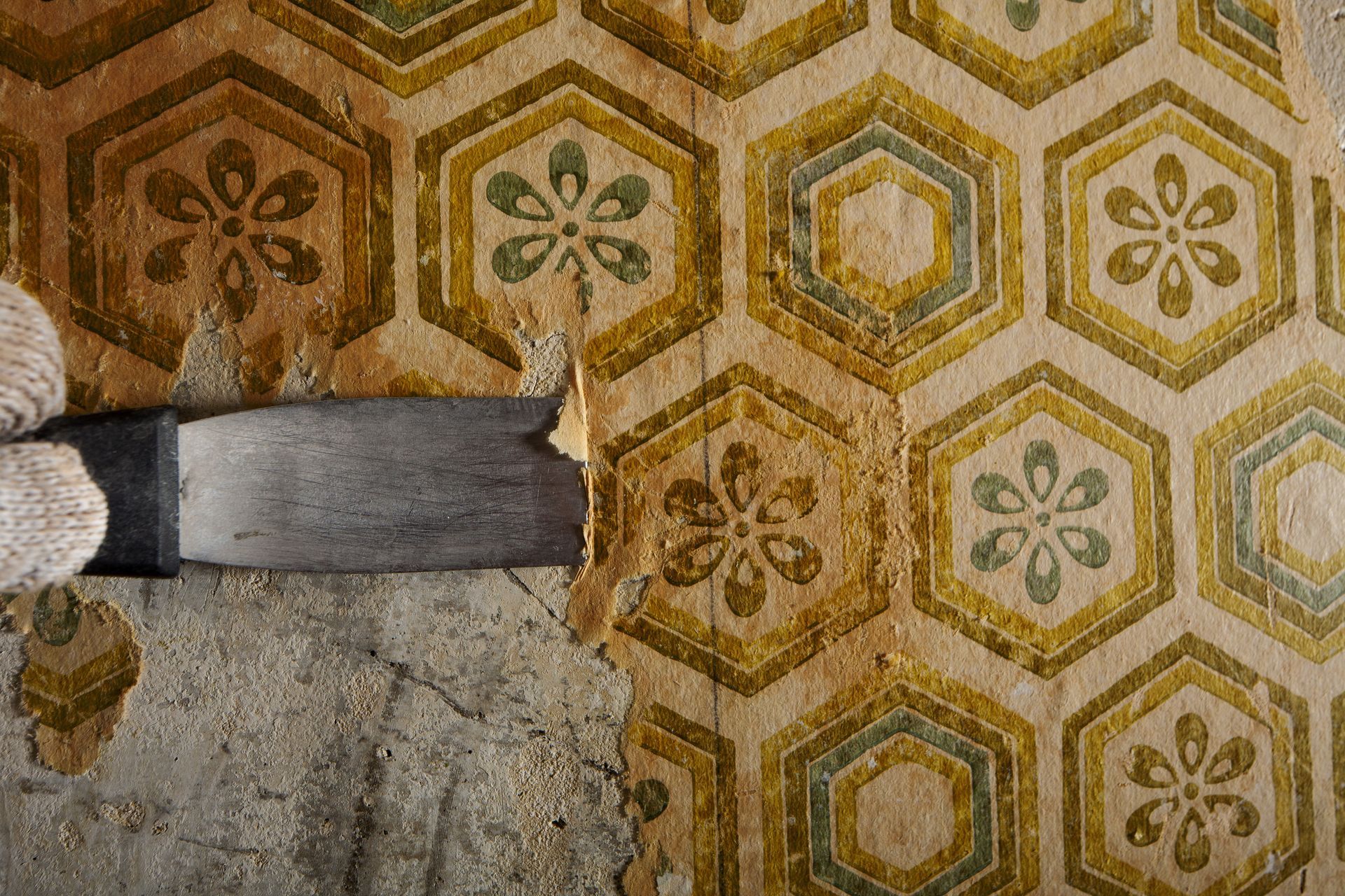 Person wearing gloves and using a scraper to remove wallpaper from the wall.