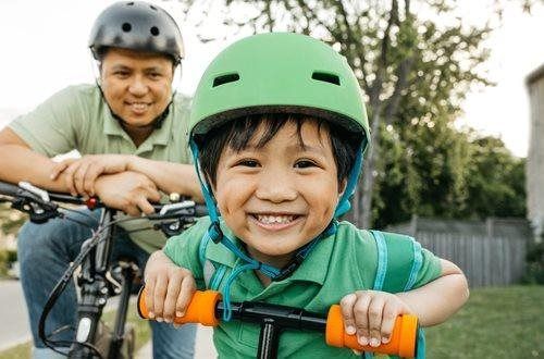 Boy in green helmet smiles while holding scooter handles; man on bicycle smiles behind him.
