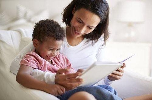 Woman and child reading a book together; both smiling and looking at the open pages.