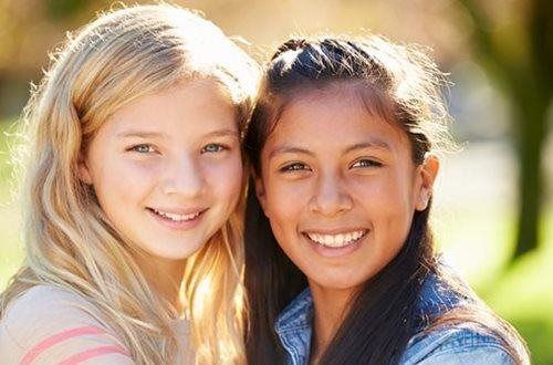 Two smiling girls with blonde and brown hair, outdoors in natural light.