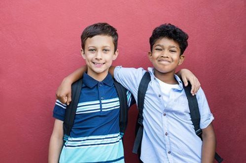 Two boys with backpacks smiling, one has his arm around the other, standing in front of a red wall.