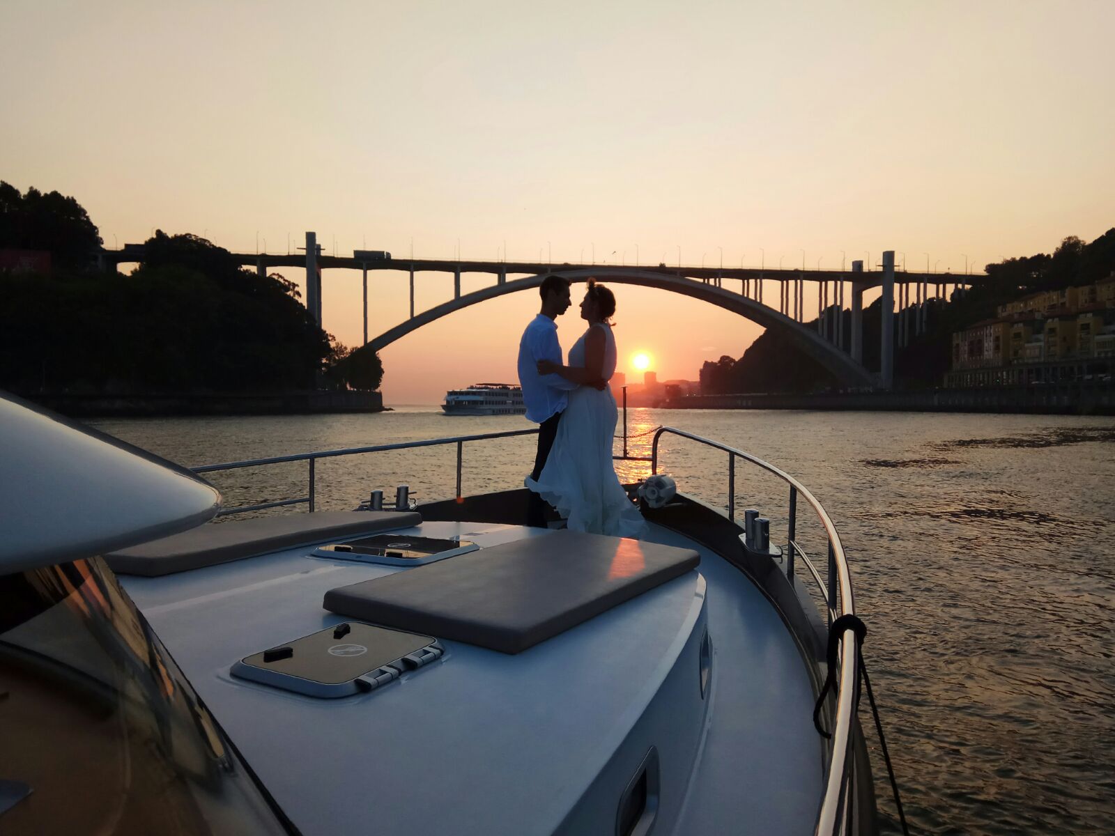 A bride and groom are standing on the bow of a boat looking at the sunset.
