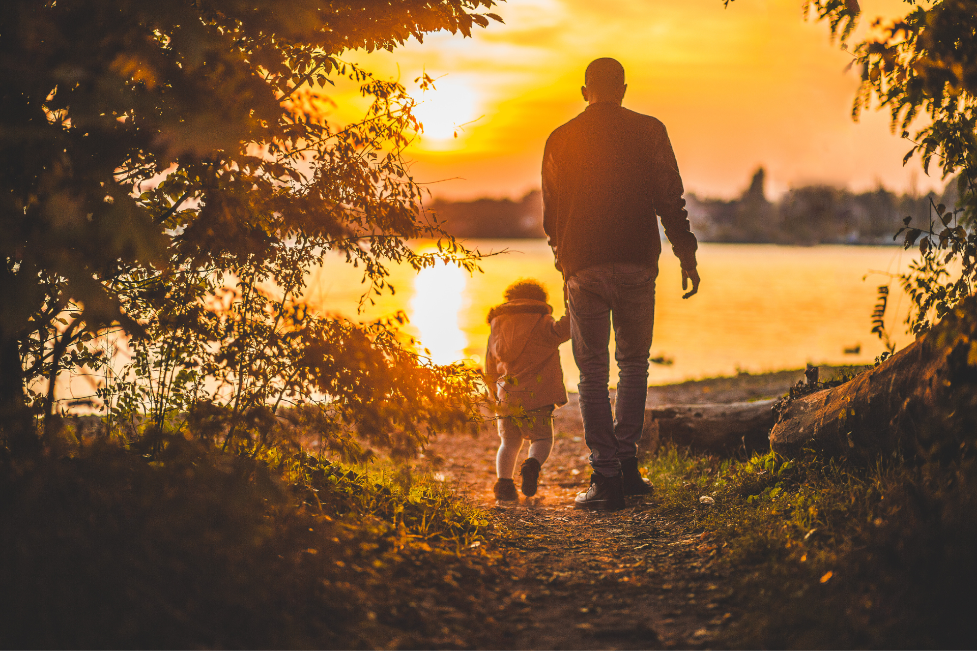 A man and a child are walking on a path near a lake at sunset.