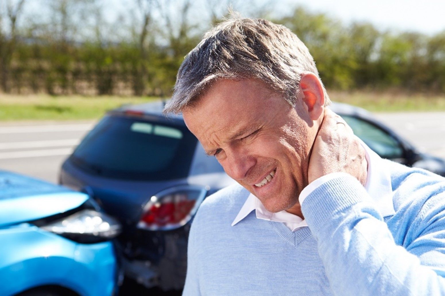 A person clutching their neck in pain with a damaged car in the background after a collision.