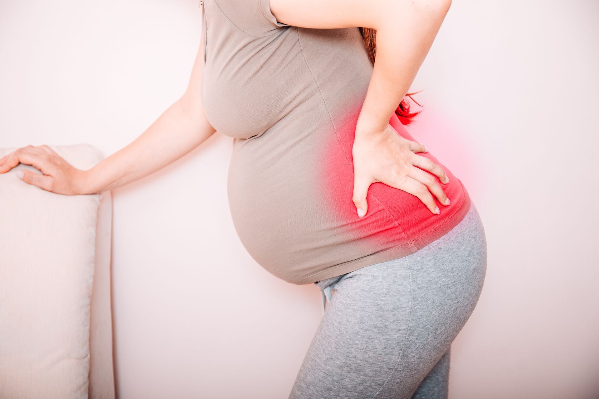 A pregnant person in a grey shirt and leggings stands, holding their lower back where a red glow indicates pain.