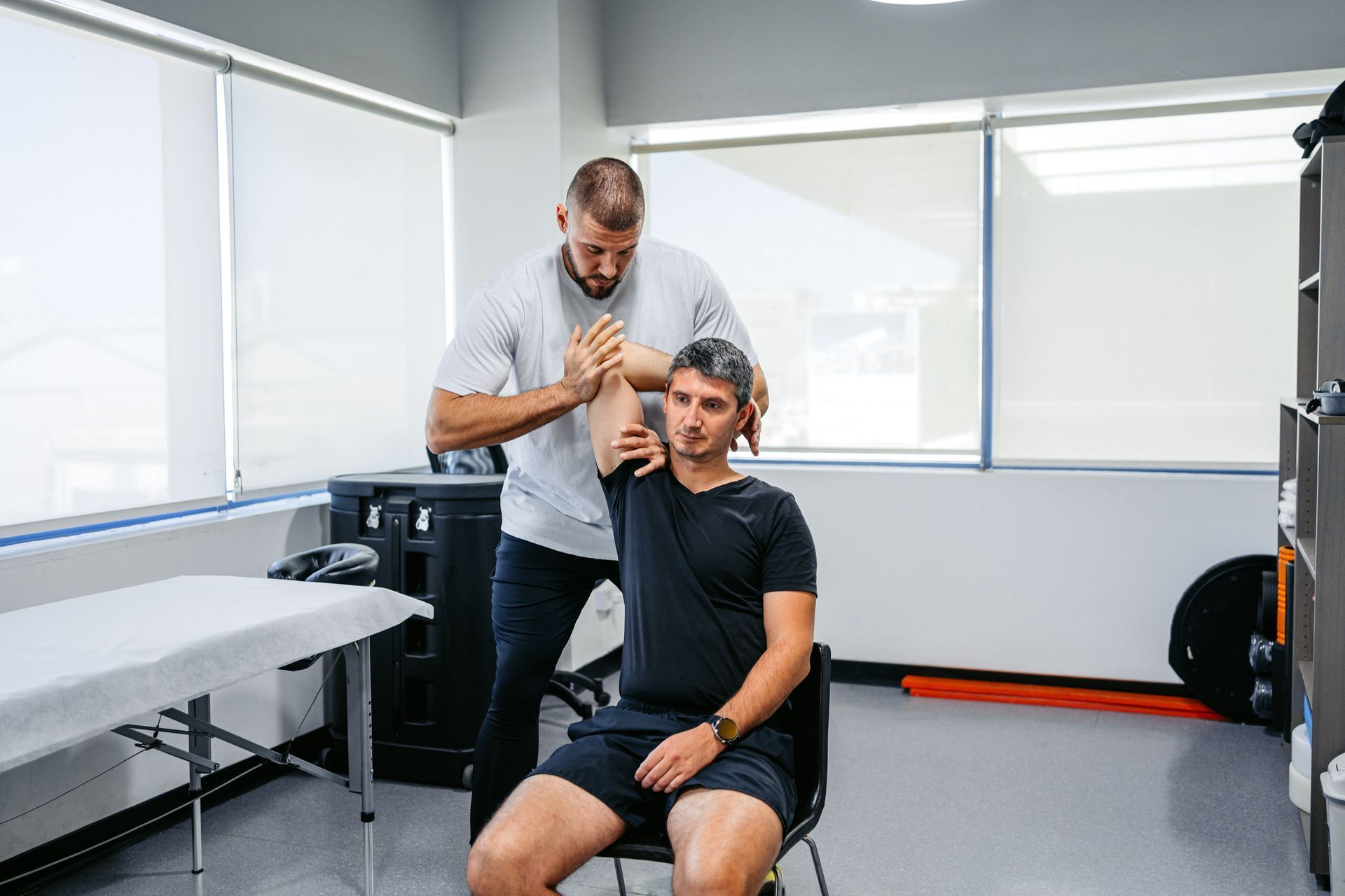 A physical therapist in a clinical room performs a shoulder assessment on a seated patient in a black t-shirt.
