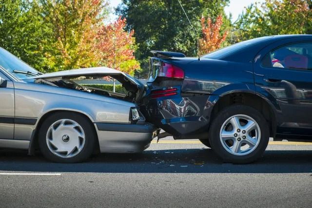 A silver car has rear-ended a dark blue car on an asphalt road with trees in the background.