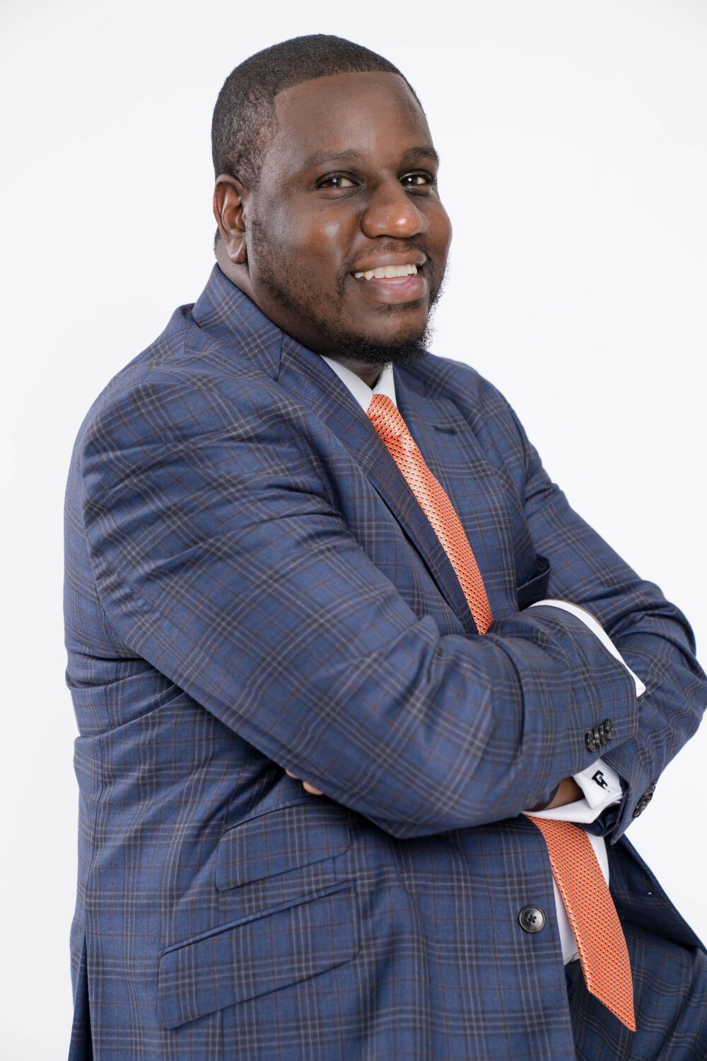 Man in blue plaid suit with arms crossed, smiling. Orange tie and white shirt. White background.