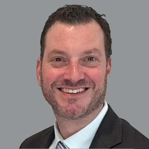 A man in a black suit jacket and shirt is looking at the camera.