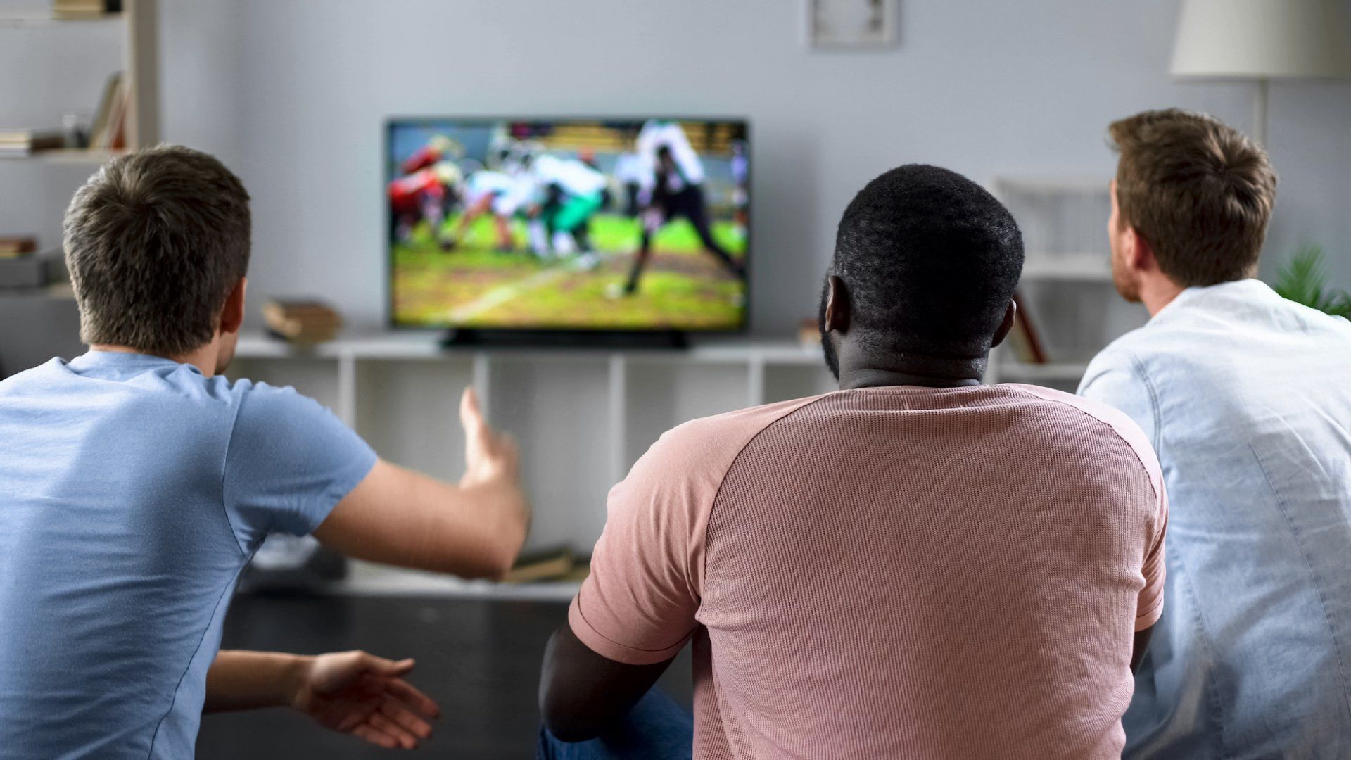 Three men are sitting on a couch watching a football game on a television.