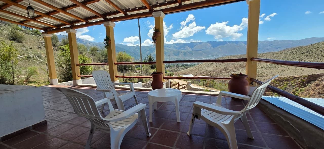 Patio con sillas blancas, mesa pequeña y macetas de barro, con vista a las montañas bajo un cielo azul.