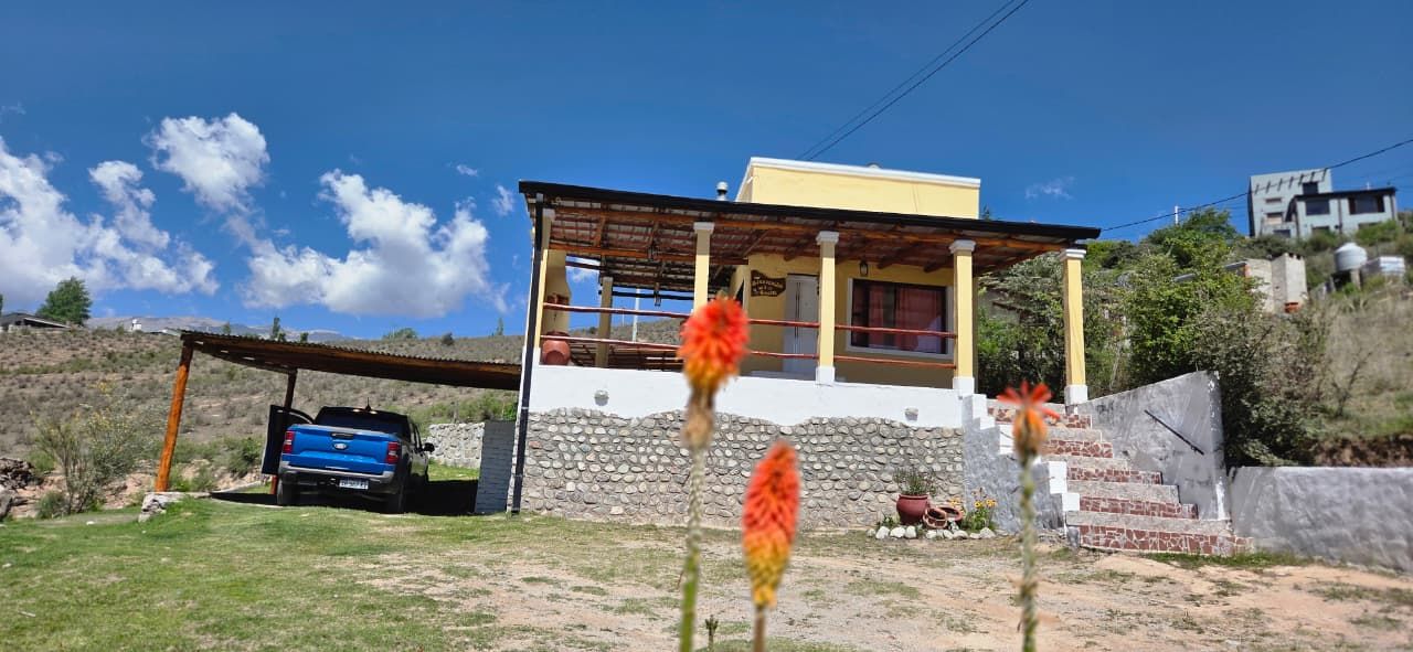 Una pequeña casa amarilla con cochera; debajo hay un camión azul aparcado. Flores rojas en primer plano.