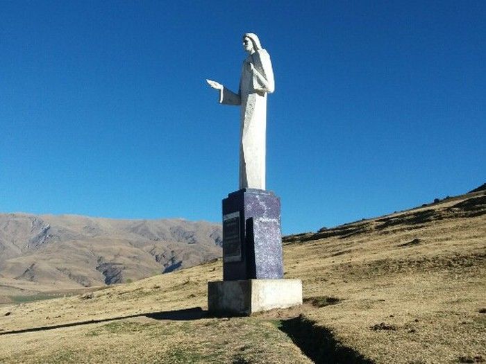Estatua de una figura con un brazo extendido sobre una base de columnas azules, frente a una colina y un cielo.