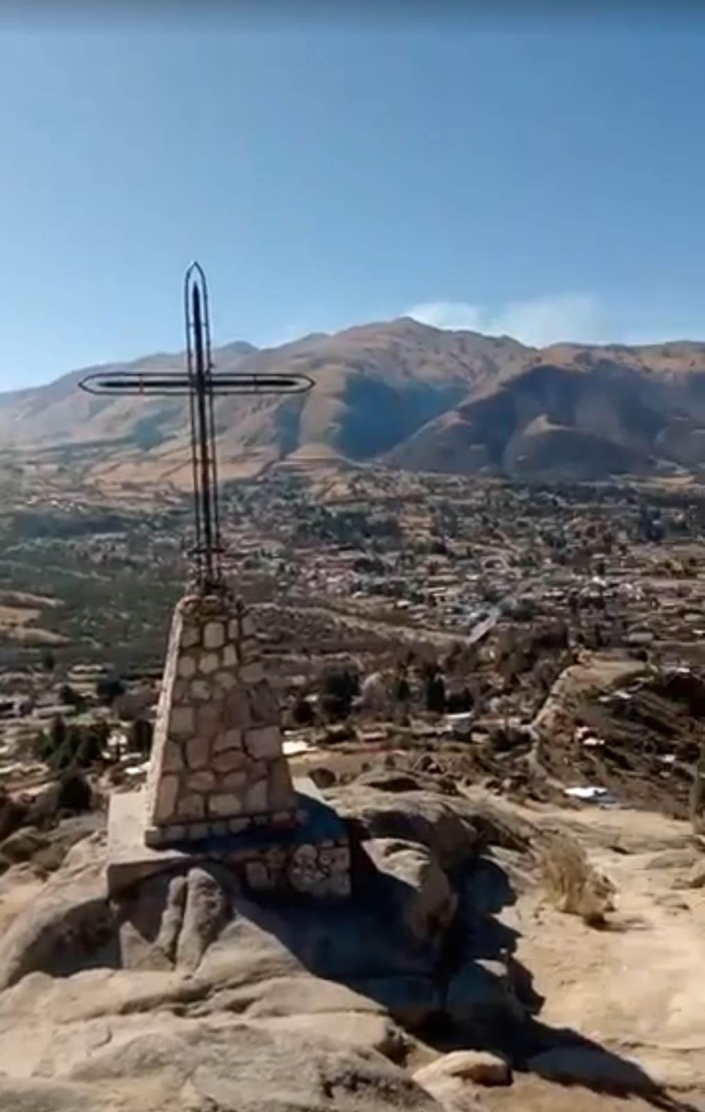 Una cruz de metal sobre un pedestal de piedra domina una ciudad y unas montañas bajo un cielo azul.