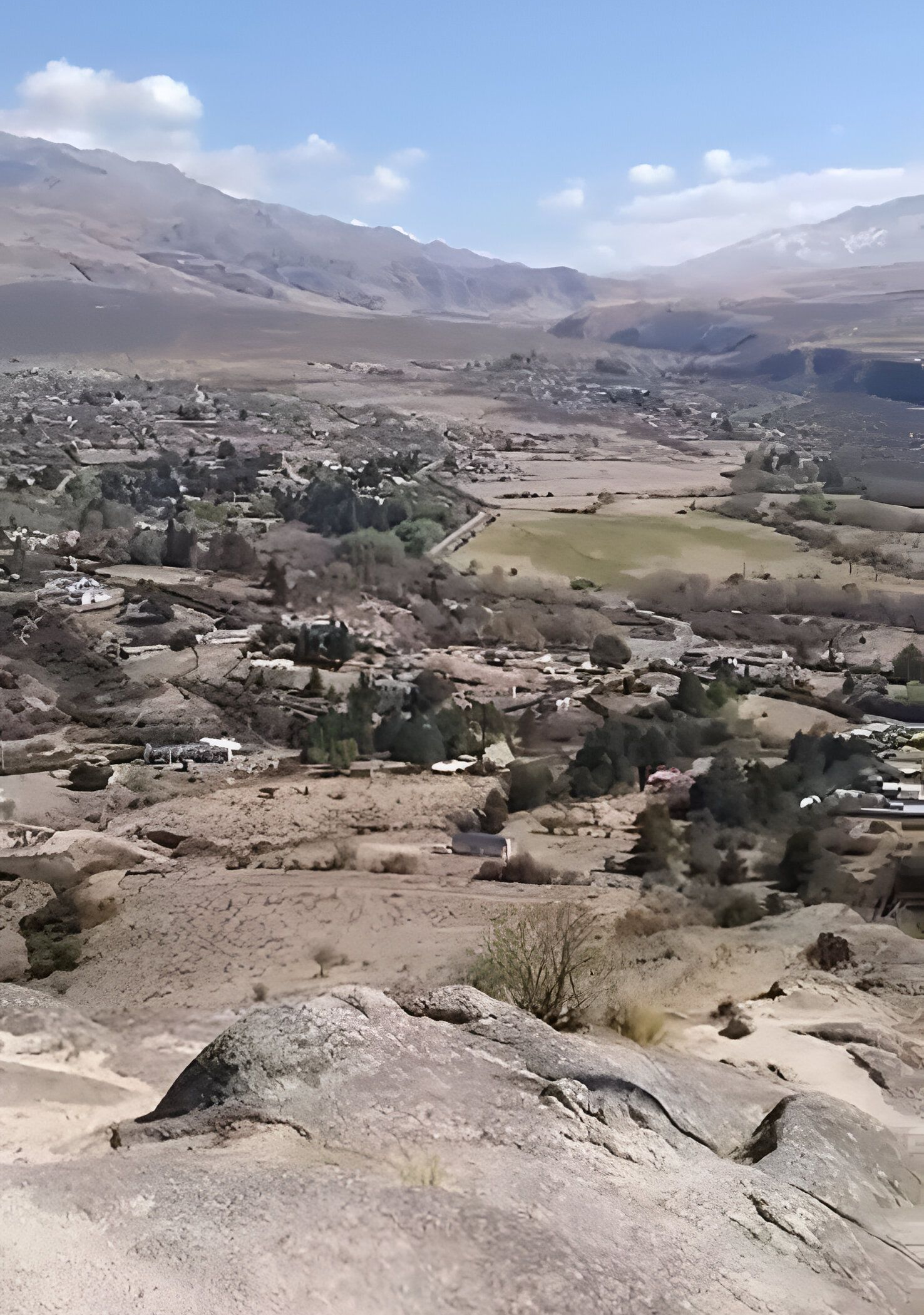 Vista del valle desde una colina rocosa; paisaje seco con edificios y montañas bajo un cielo azul.