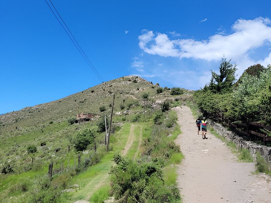 Un camino de tierra asciende por una ladera verde bajo un cielo azul, con dos personas caminando cuesta arriba.