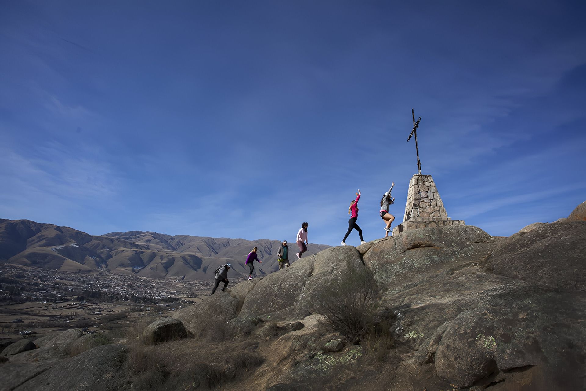 Gente caminando hacia un marcador de piedra con una cruz en lo alto de un pico rocoso; cielo azul de fondo.