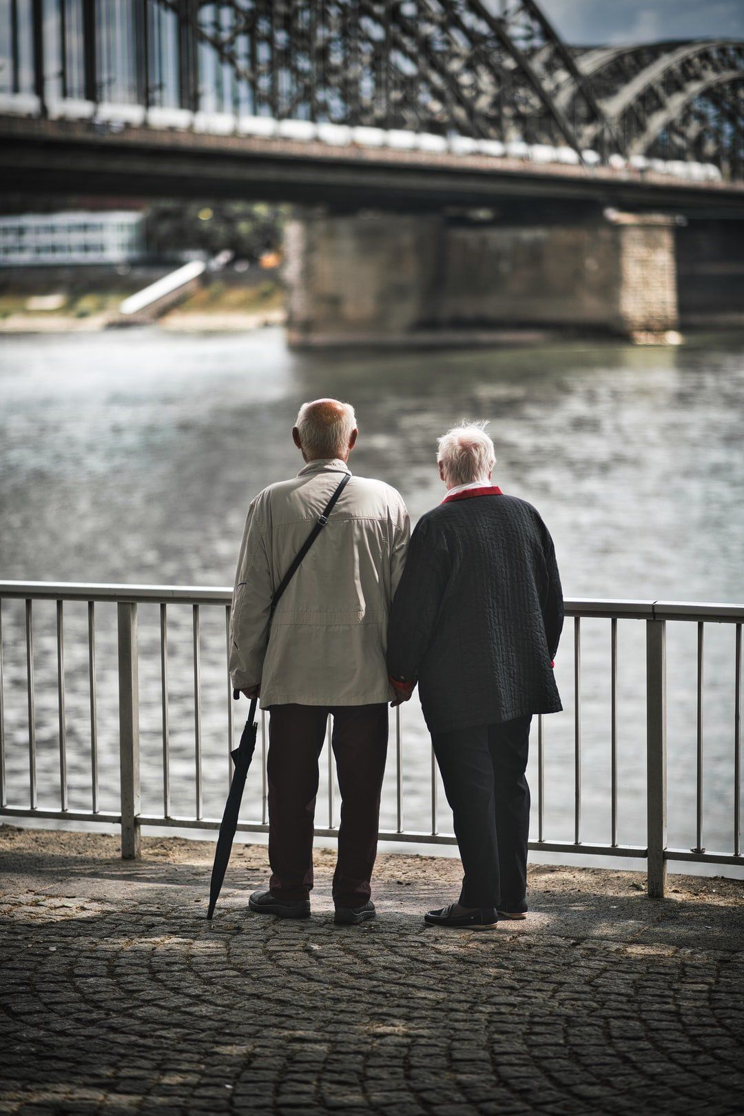 Two people, holding hands, stand by a river, looking toward a bridge.