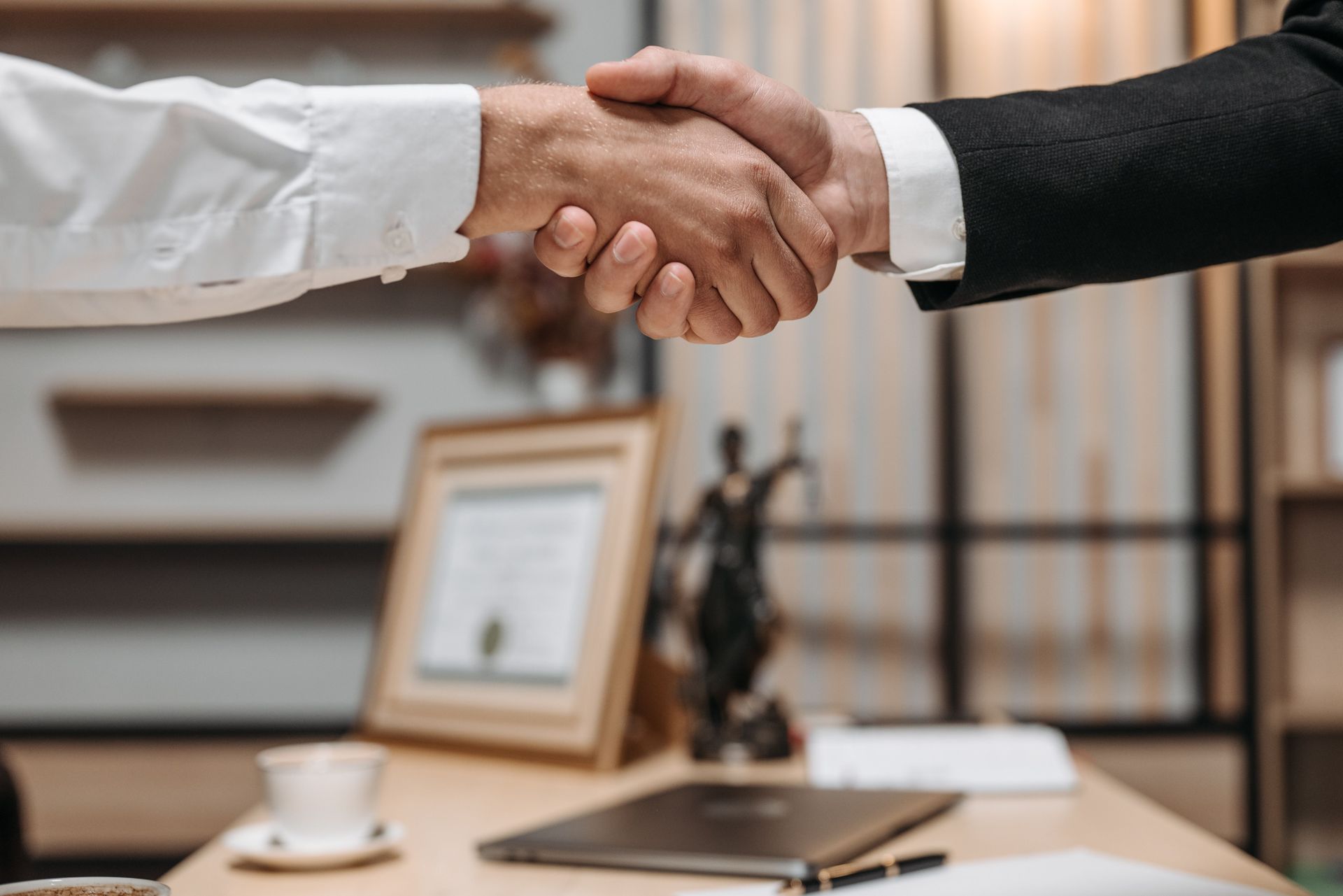 Handshake over an office desk with framed document and coffee cup in the background