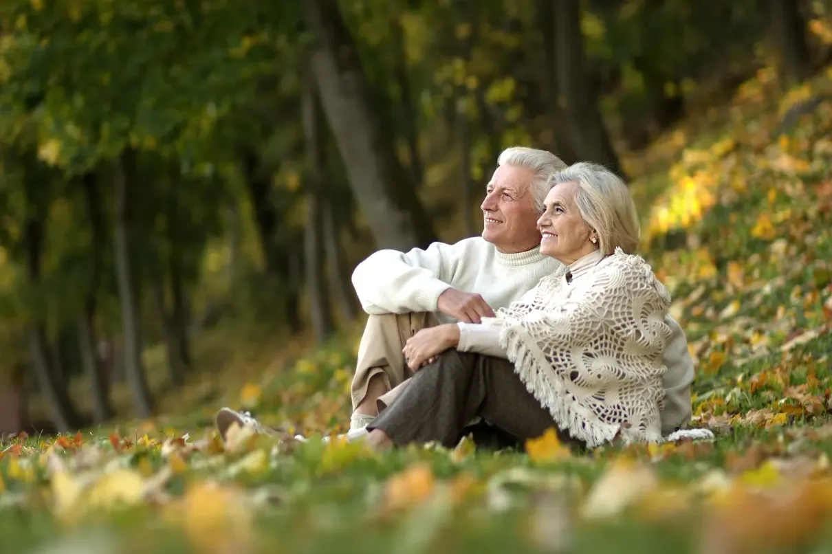 Two people sitting on a grassy hillside covered in autumn leaves, smiling while looking toward the distance.