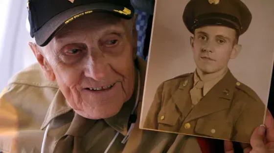 An elderly veteran wearing a military cap smiles while holding a vintage black-and-white photo of himself in uniform.