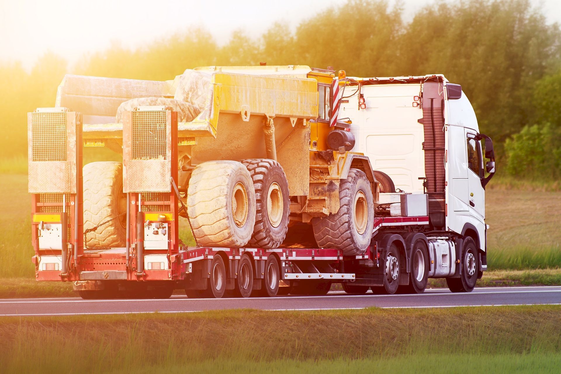 A heavy industrial truck with a trailer platform hauls a construction truck on a highway road.