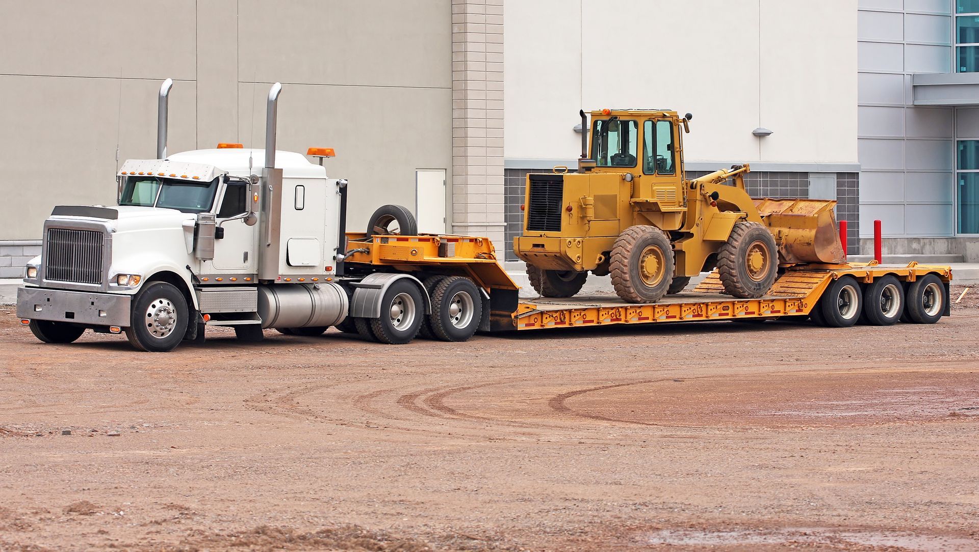 A white semi-truck hauling a yellow bulldozer on a flatbed trailer in an industrial area.