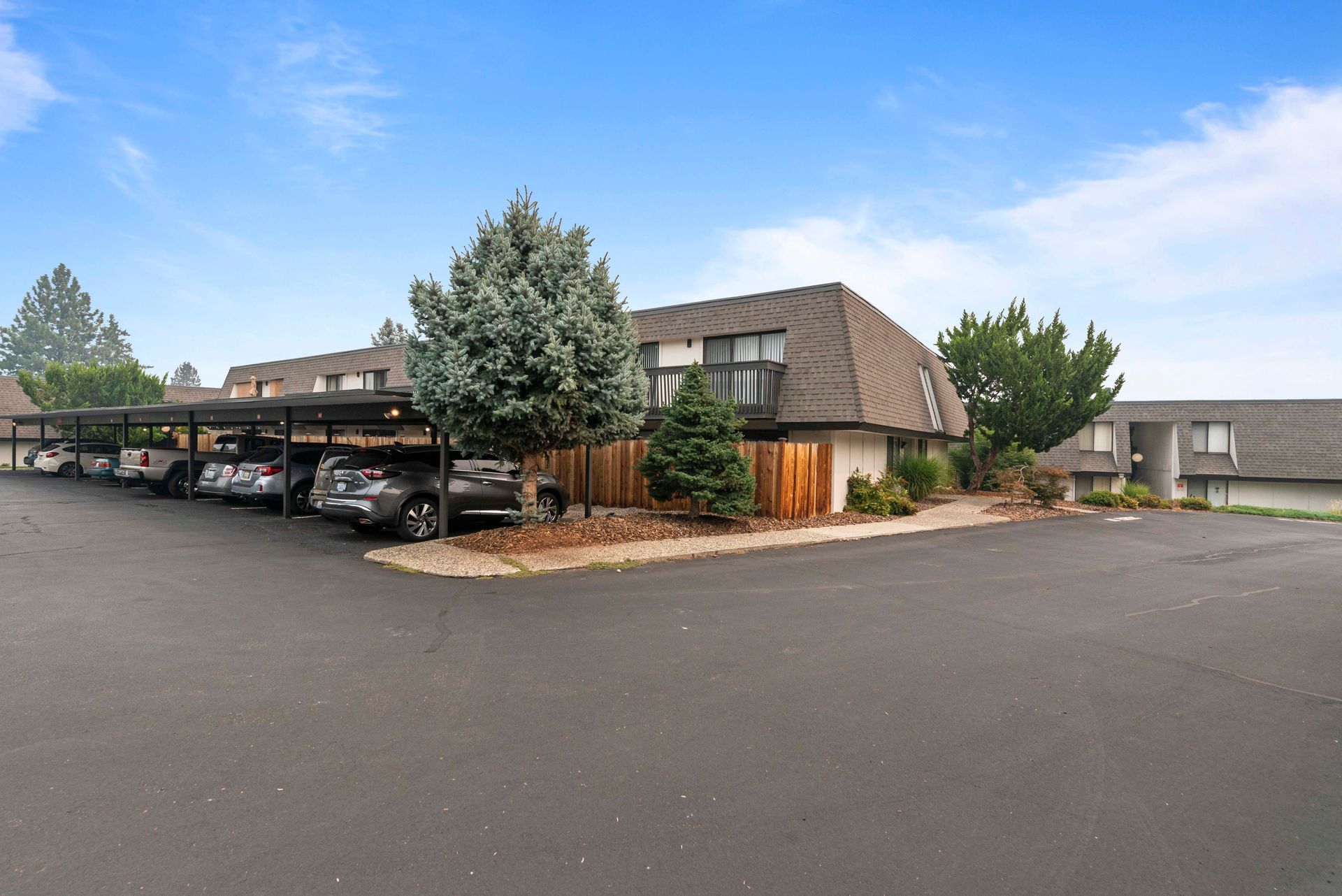 A row of cars are parked under a covered parking lot in front of a house.