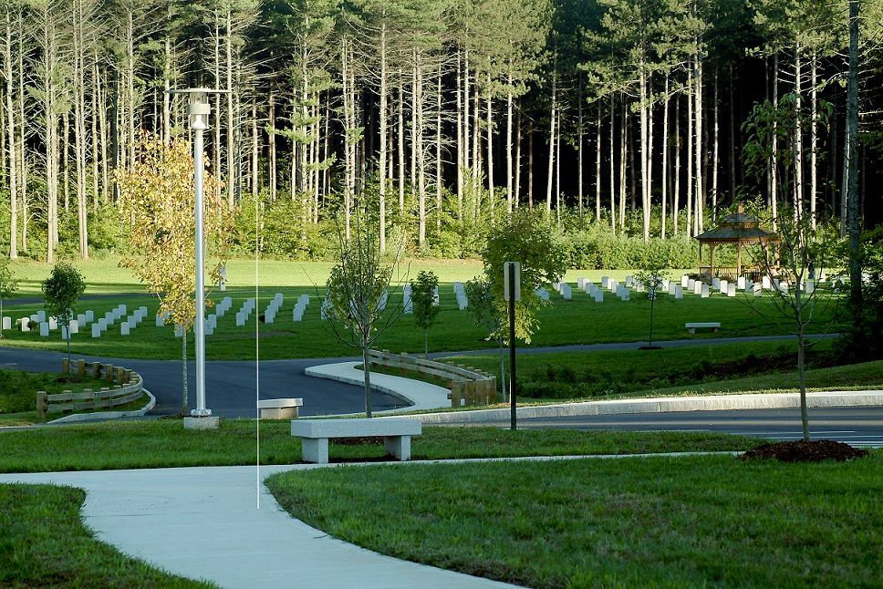 A paved path winds through a cemetery with rows of headstones, trees, and a gazebo in the background.