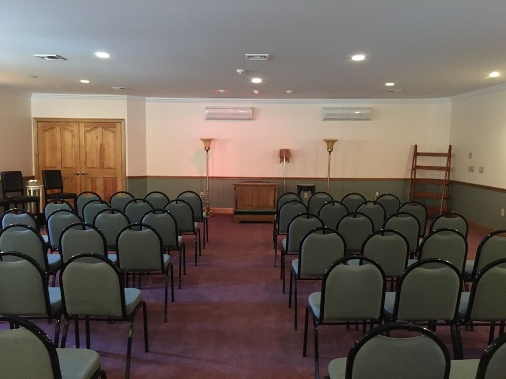 Rows of chairs face a wooden cabinet in a beige-walled room, likely a funeral home.