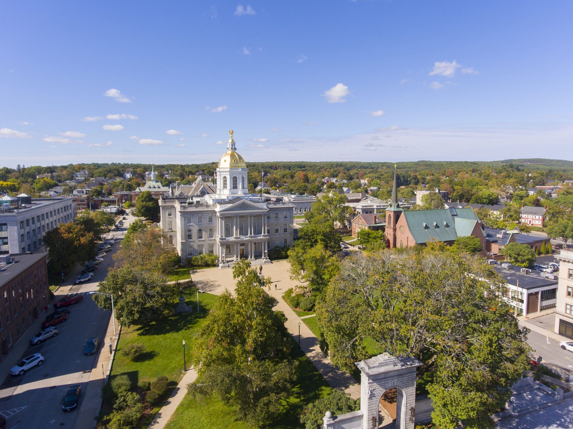 Aerial view of the New Hampshire State House in Concord with a gold dome, surrounded by trees and buildings on a sunny day.