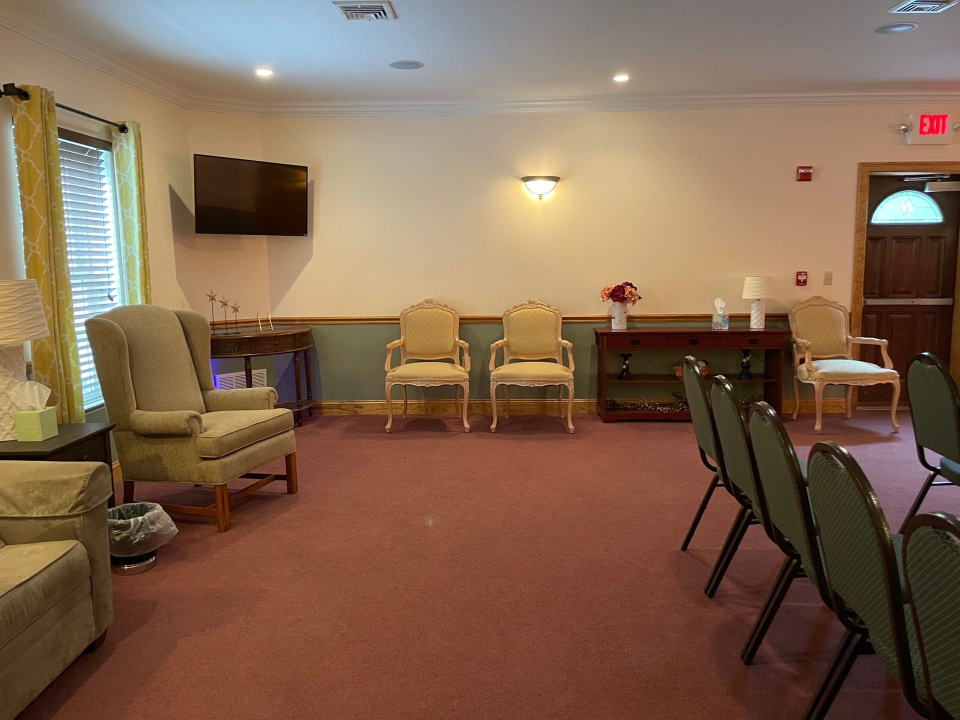 Funeral home interior with chairs, TV, and doorway. Pink carpet, beige walls.