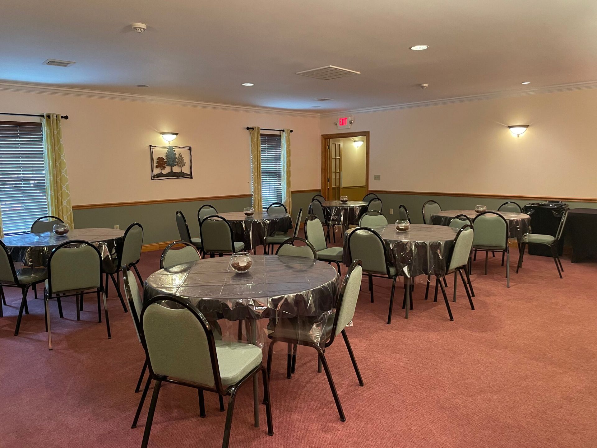 A room with round tables and chairs, ready for a gathering. Pink carpet, light walls, and a doorway.