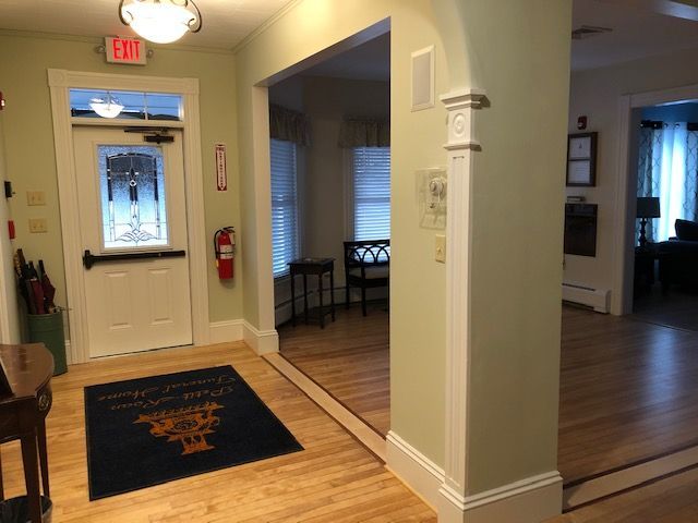Entryway with a door, fire extinguisher, and welcome mat. Light green walls, wood floors, and a glimpse into another room.
