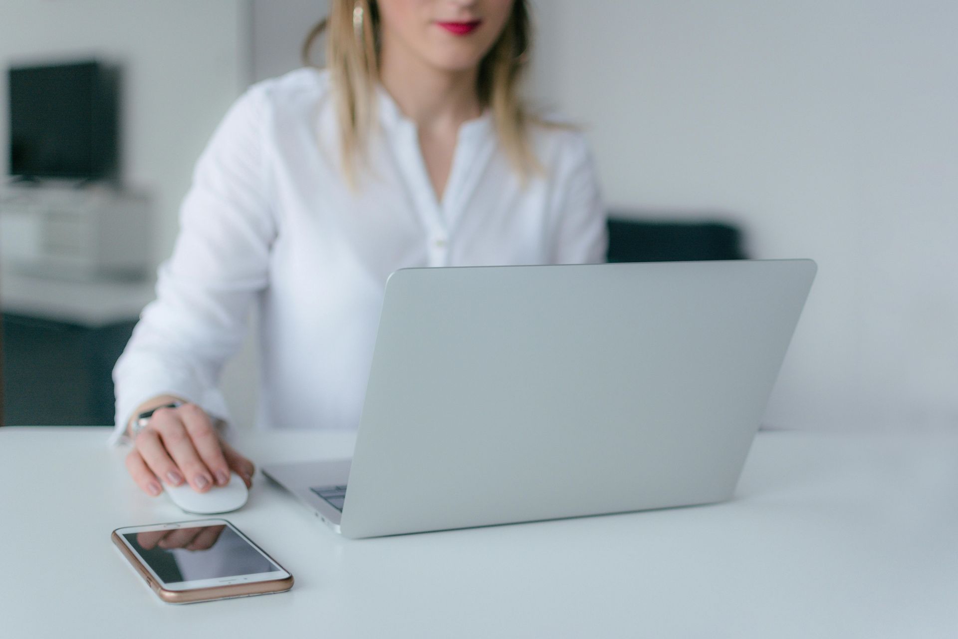 Woman using a laptop and mouse at a desk, with a smartphone in front.