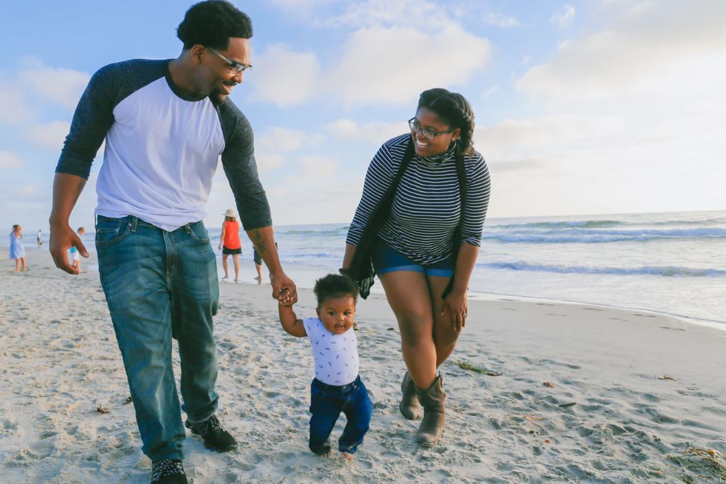 Happy family walking on a beach, helping their toddler walk