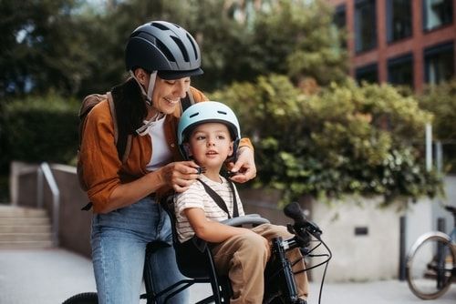 Mother fastening sons' bike helmet on head, carring him on child bike carrier, seat. Mom commuting with a young child through the city on a bicycle.