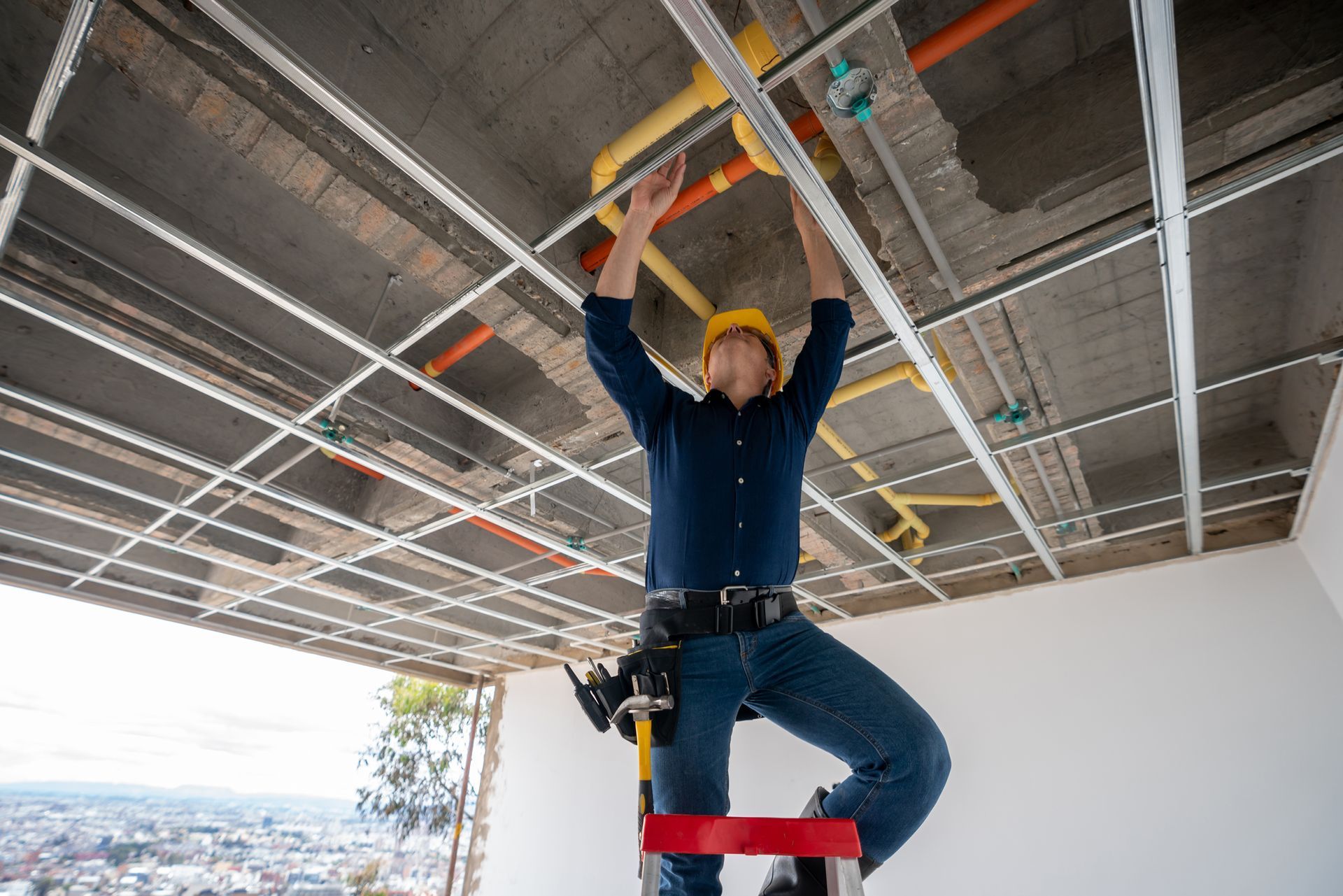 Construction worker in a yellow hard hat installs ceiling grid while standing on a red step ladder; exposed pipes and wires visible.