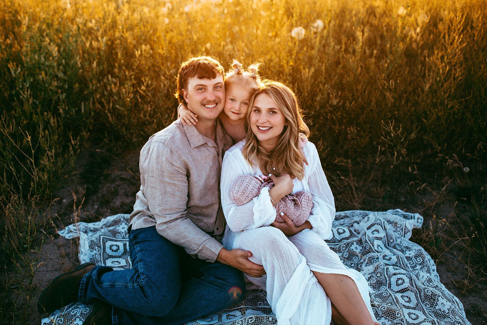 Family of four smiles for the camera while sitting on a blanket in a field at sunset. Family of four smiles for the camera while sitting on a blanket in a field at sunset.