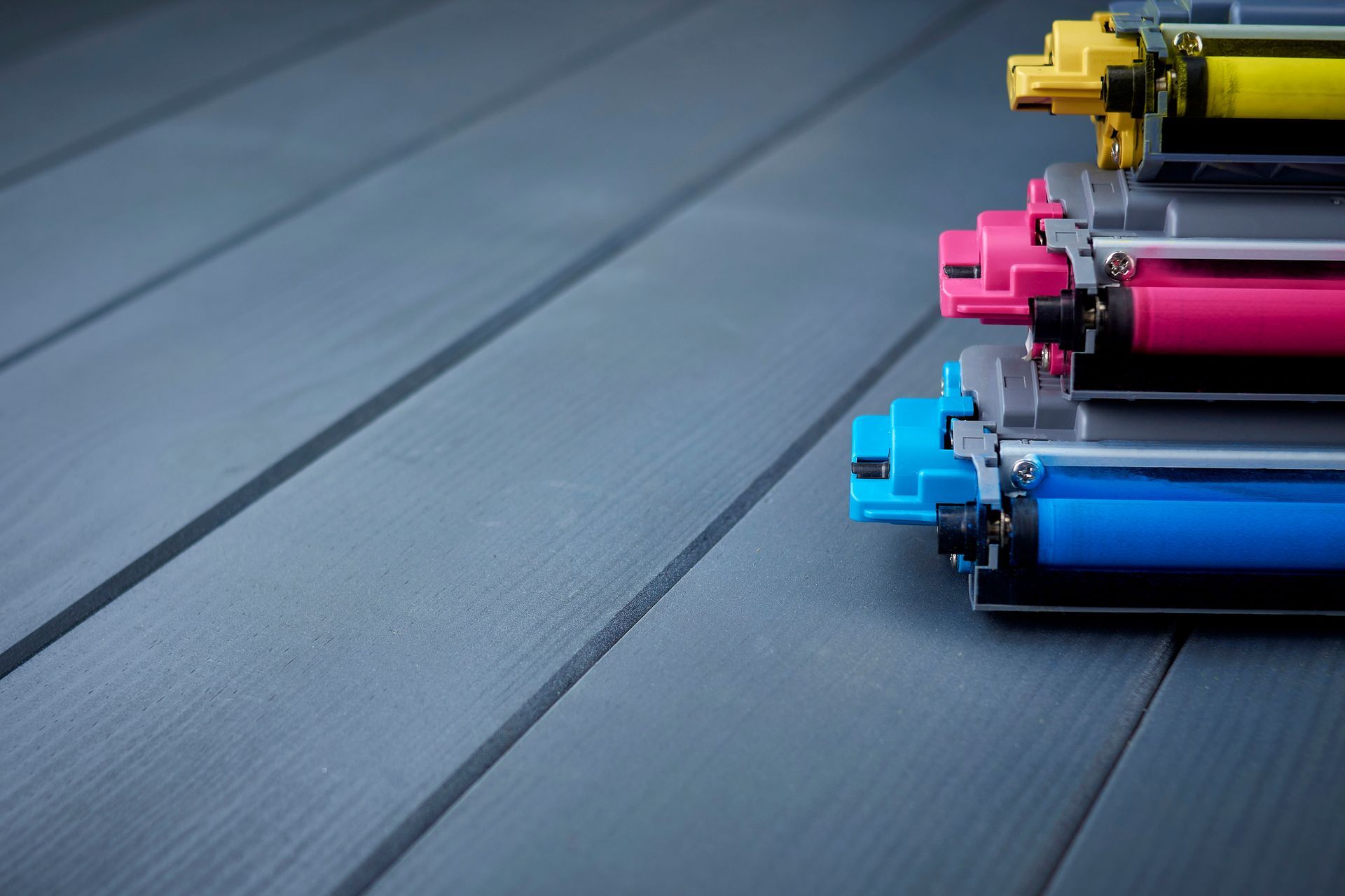 Cartucce di toner ciano, magenta, giallo e nero impilate su una superficie di legno grigio.