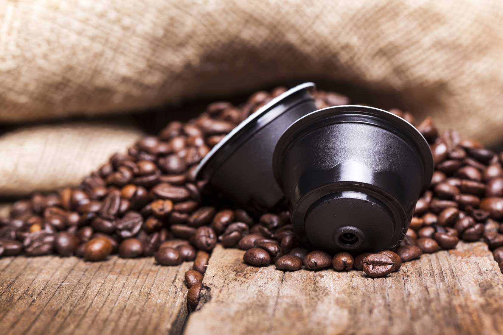 Capsule di caffè su un letto di chicchi di caffè tostati, disposte su una superficie di legno con tela di iuta sullo sfondo.