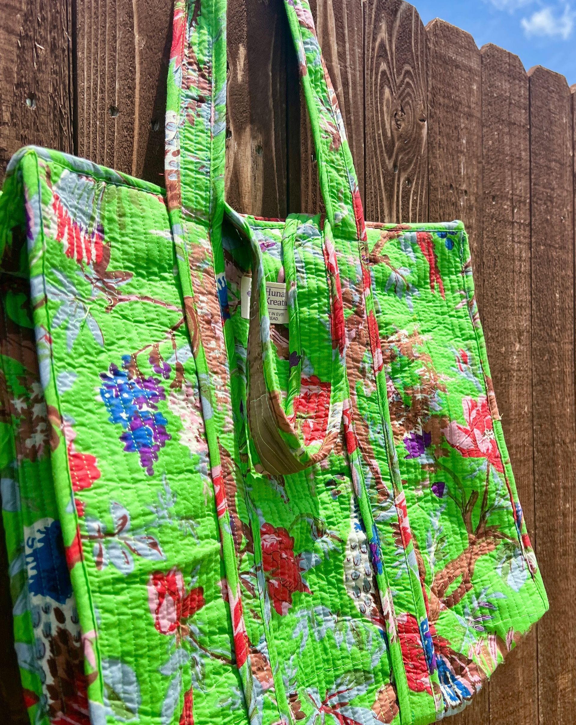 A green quilted tote bag is hanging on a wooden fence.