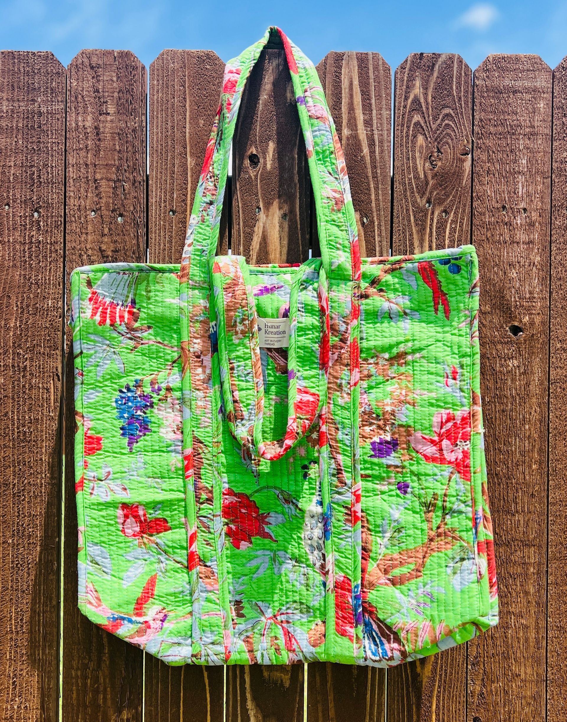 A green quilted tote bag is hanging on a wooden fence.